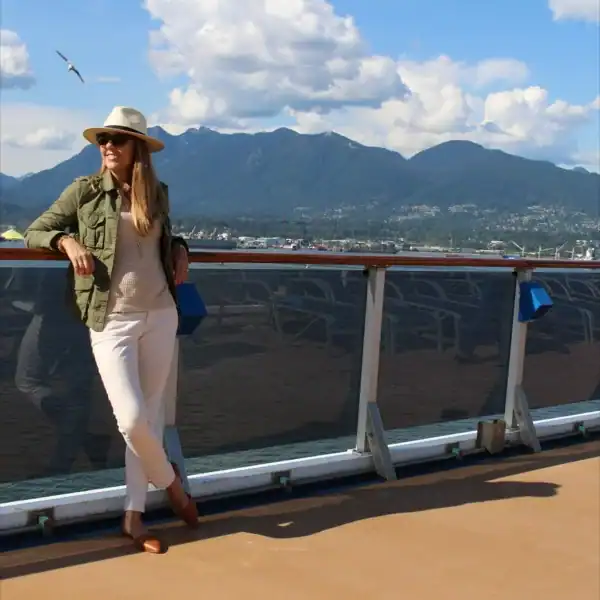 woman in casual outfit and hat leaning on a cruise railing with Alaska mountains in the background for the post on Alaska cruise packing list by month