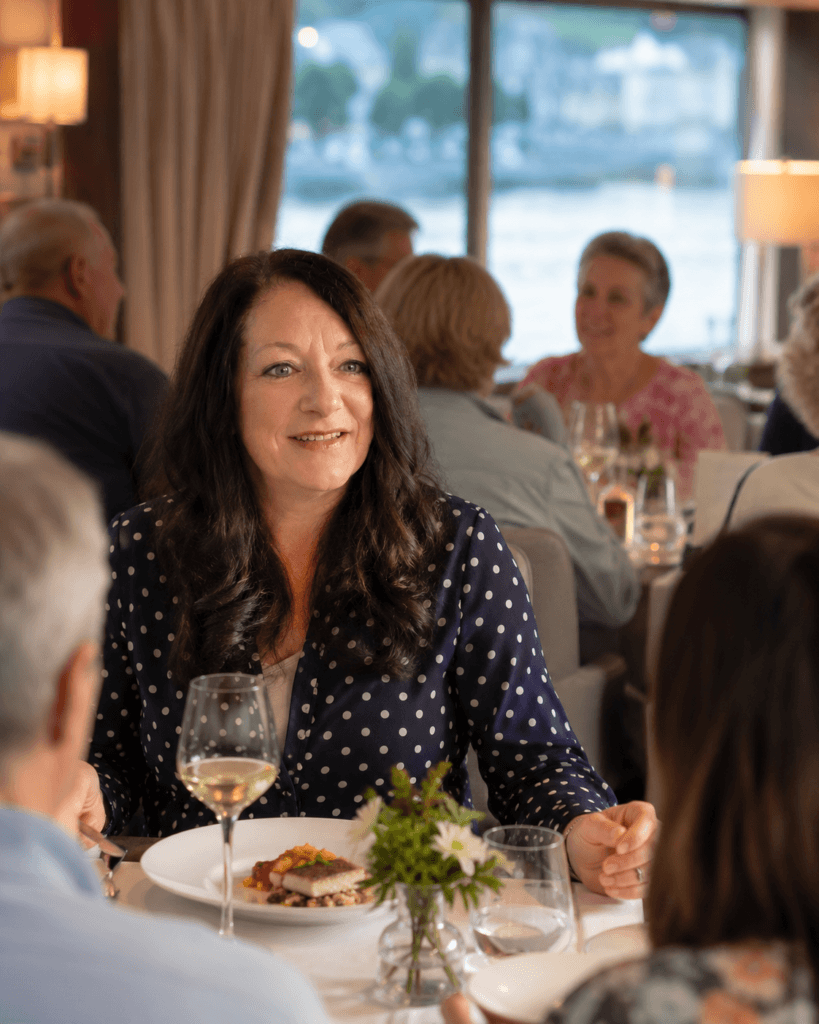 Solo traveler sitting at a communal dining table on a European river cruise ship, enjoying dinner with fellow passengers. 